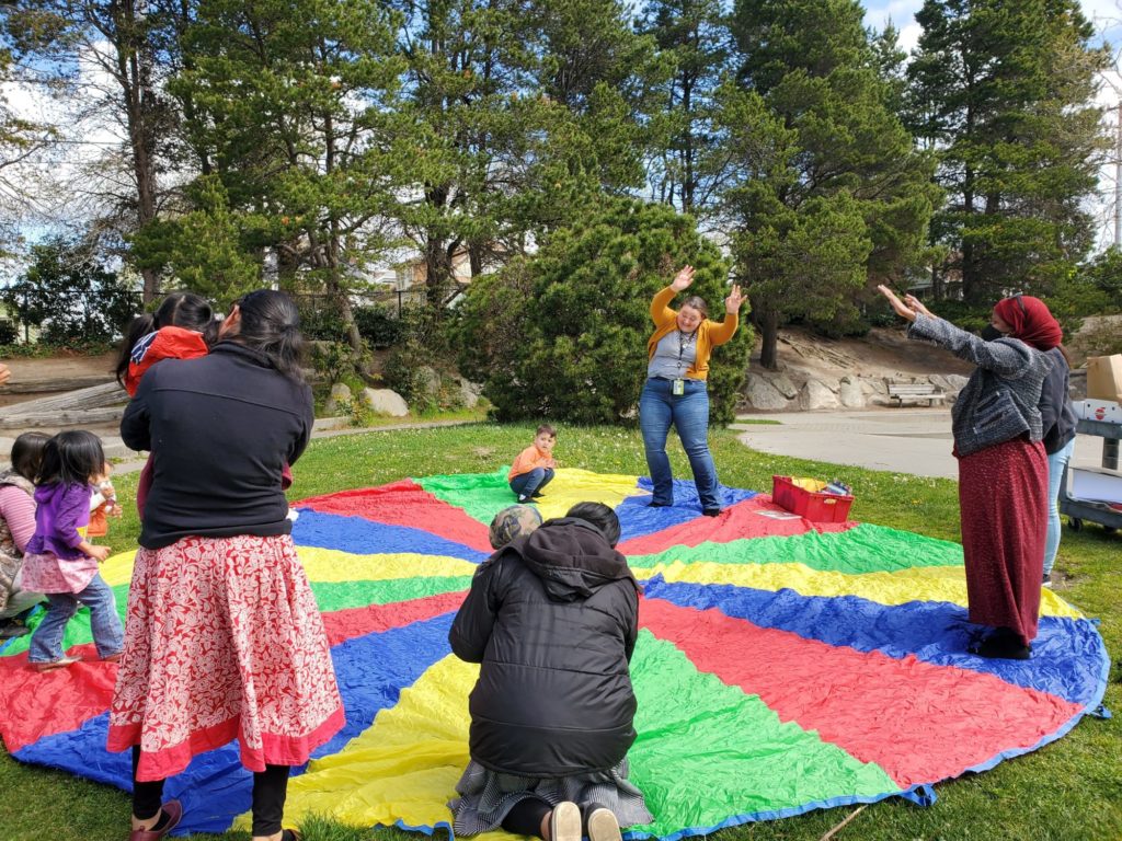 Children and their caregivers stand in a circle around a play parachute, some with arms raised, taking part in an activity.