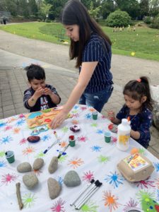 Two kids sit at an activity table at a park with an adult standing between assisting them as they paint and decorate rocks.