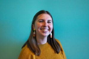 A headshot of Community Connector Nicole Freitas (she/her) in front of a turquoise background.